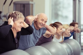 Elderly couple doing pilates class at the gym with a group of diverse younger people balancing on the gym ball with raised arms to tone their muscles in an active retirement concept.jpeg